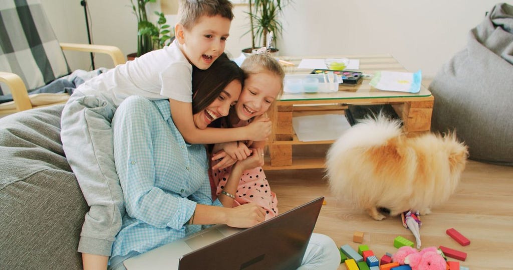 Mom on floor with laptop smiling while boy and girl hug her