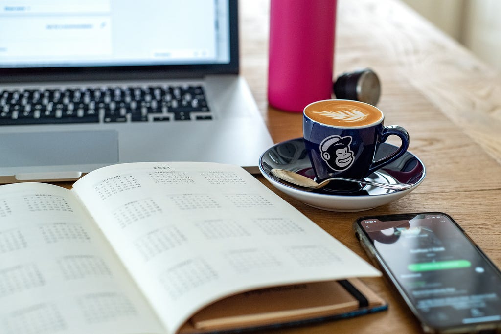 A laptop, cell phone, calendar, and a coffee on a desk.