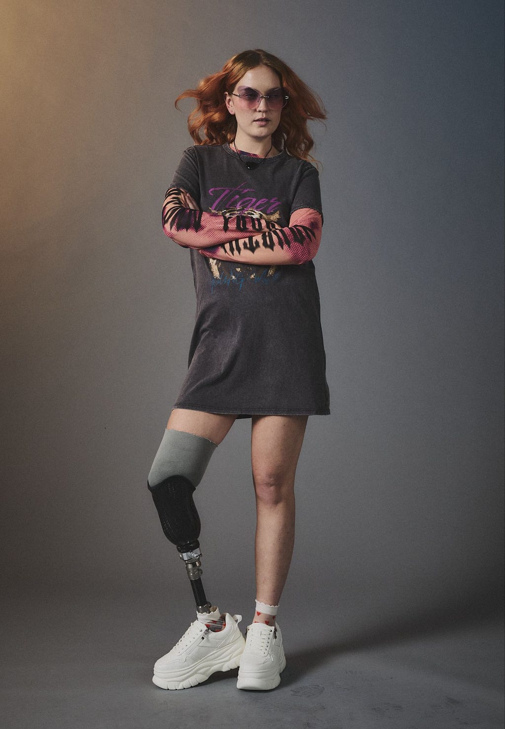A white person with red hair and a prosthetic leg, wearing a short, grey t-shirt dress,  chunky white sneakers and frameless sunglasses with pink lenses, pictured against a grey gradient background with their arms crossed.