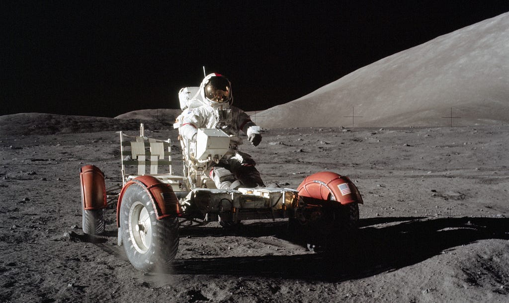 An astronaut drives a lunar rover across the rocky surface of the Moon, with hills in the background and a dark, starless sky above.