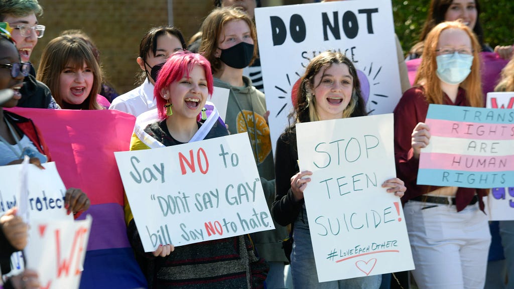 Young people holding signs at a protest that read “say no to ‘don’t say gay’ bill, say no to hate”; “stop teen suicide”; “trans rights are human rights.” One person holds a bisexual pride flag; another has a nonbinary pride flag wrapped around their shoulders.