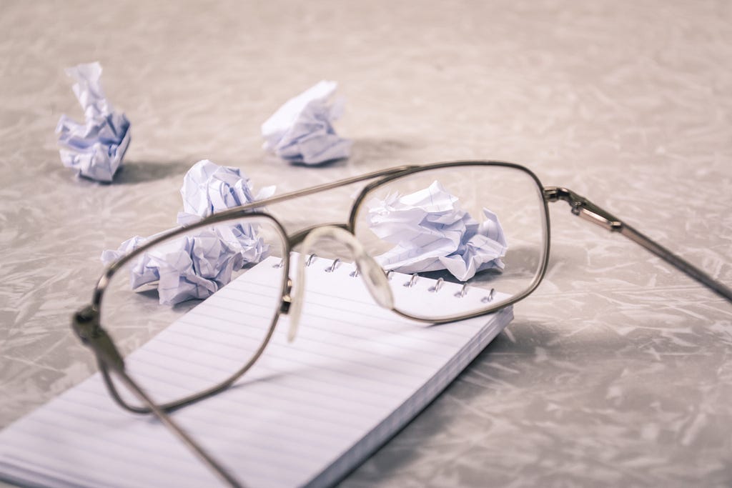 A pair of glasses on top of a blank notebook with some crumbled papers on the side