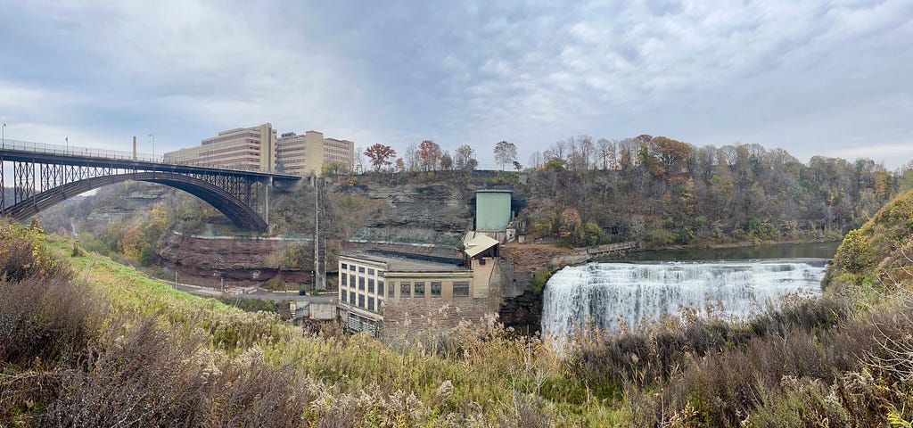 panorama view of the Driving Park Avenue Bridge, office buildings, a river bed, a waterfall, and vegetation along the Genesee River.