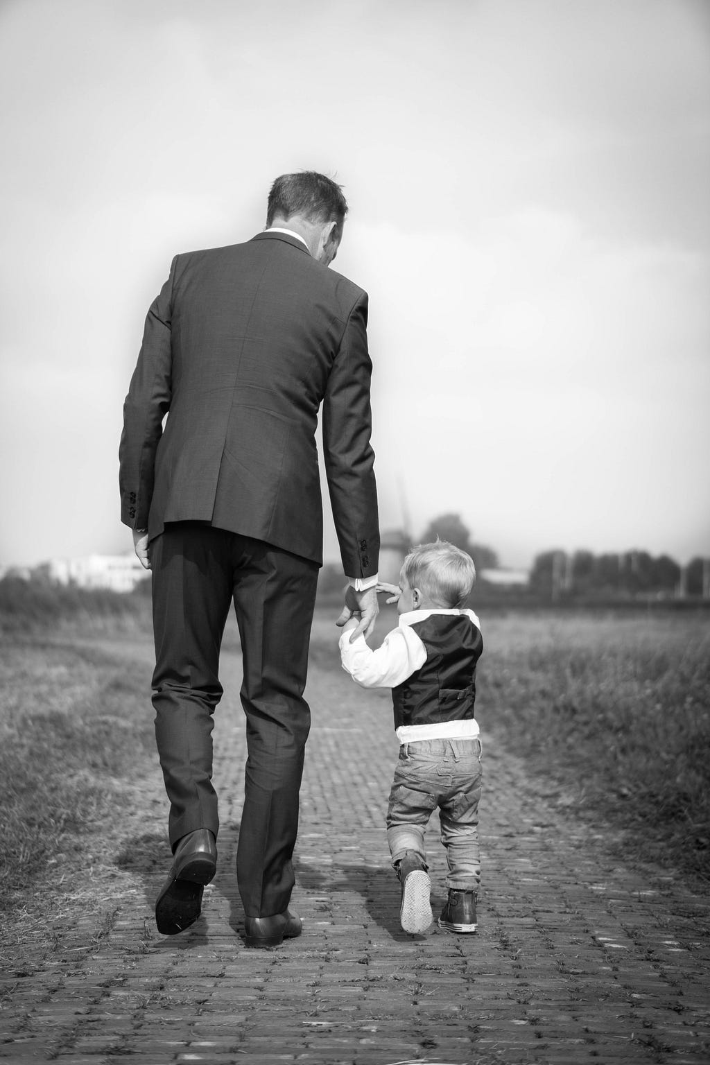An adult man, wearing a suit, is seen walking down a path, away from the camera. His right hand is reaching downwards, to let the toddler to his right grab hold of it and walk with him.