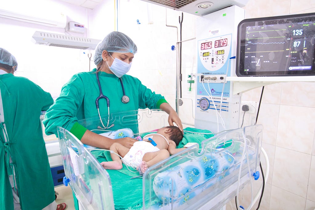 A health worker looks down at a baby in a child-sized hospital bed surrounded by medical monitors and equipment.