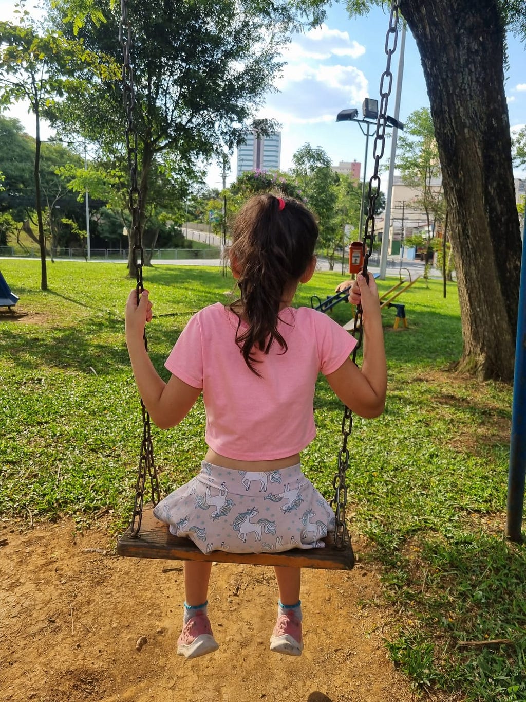 Menina branca de cabelos castanhos amarrados. Usando blusa rosa e shorts com unicórnios estampados. Sentada, de costas, em um balanço de um parquinho de rua.