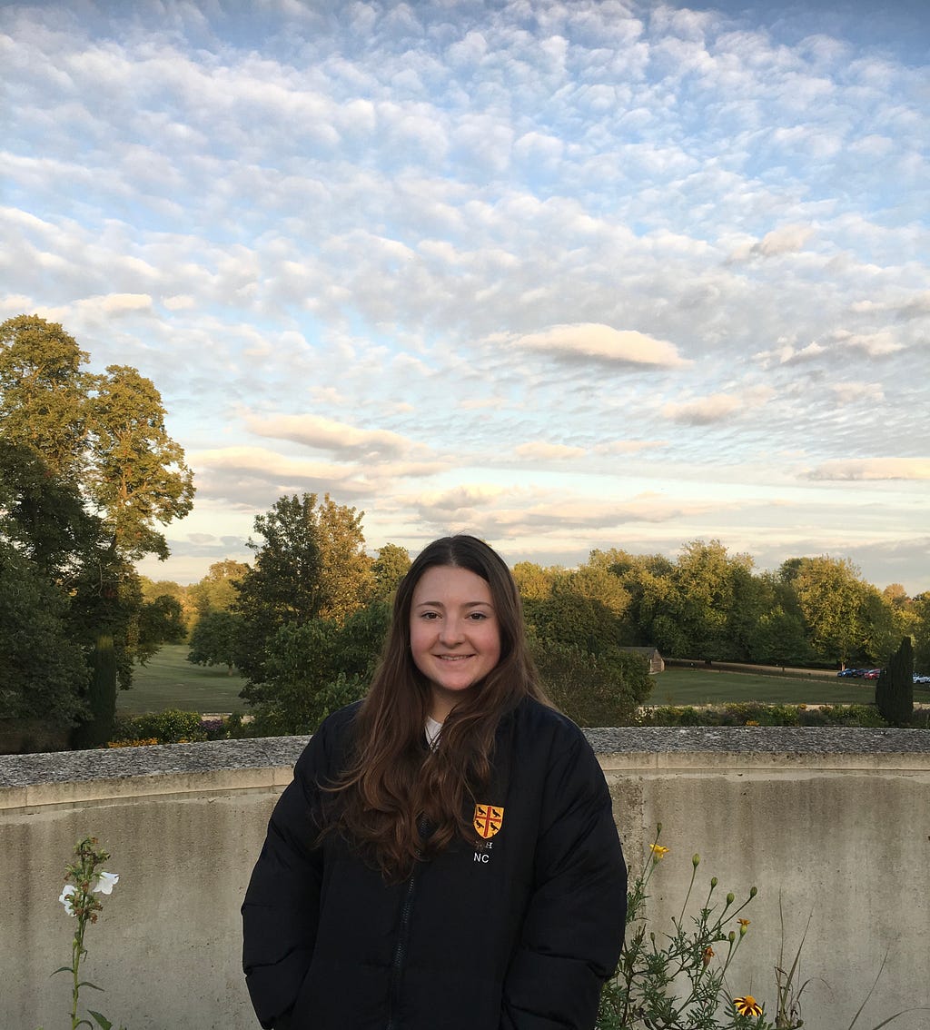 A photograph of Natasha, who is smiling in front of a picturesque country landscape, with a pond, blue skies and green trees in the background.