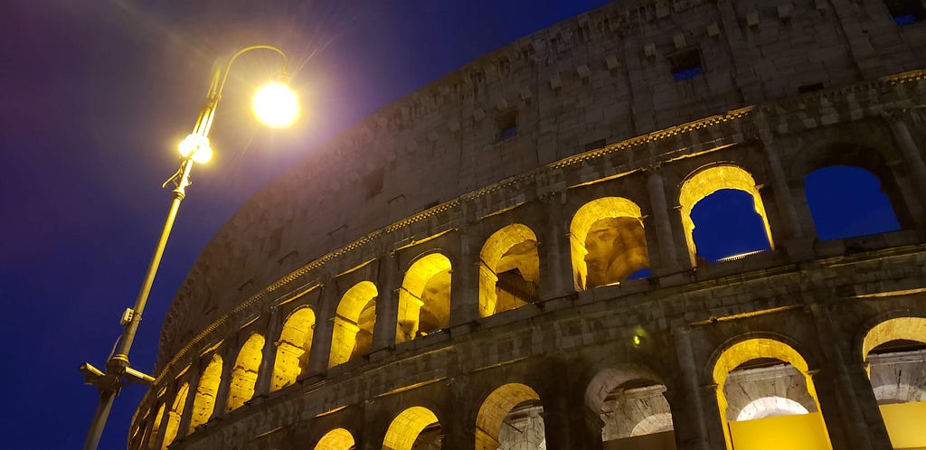 A night-time image of the Roman Colosseum with a bright lamp post glaring down from above. The Colosseum is lit from behind.