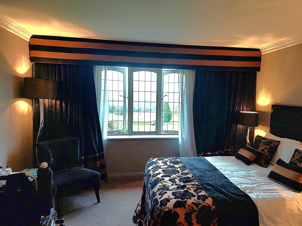 A photo of the windows in a hotel room, framed with heavy curtains. The route to the windows and the curtains is blocked by the bed and a heavy chair.