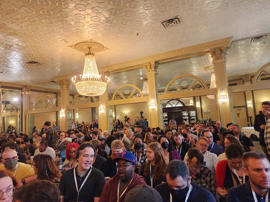 [ID: A room in the Austin Club packed with screenwriters of various genders and skin tones. The room has a white ceiling, cream colored arches, and a chandelier.]