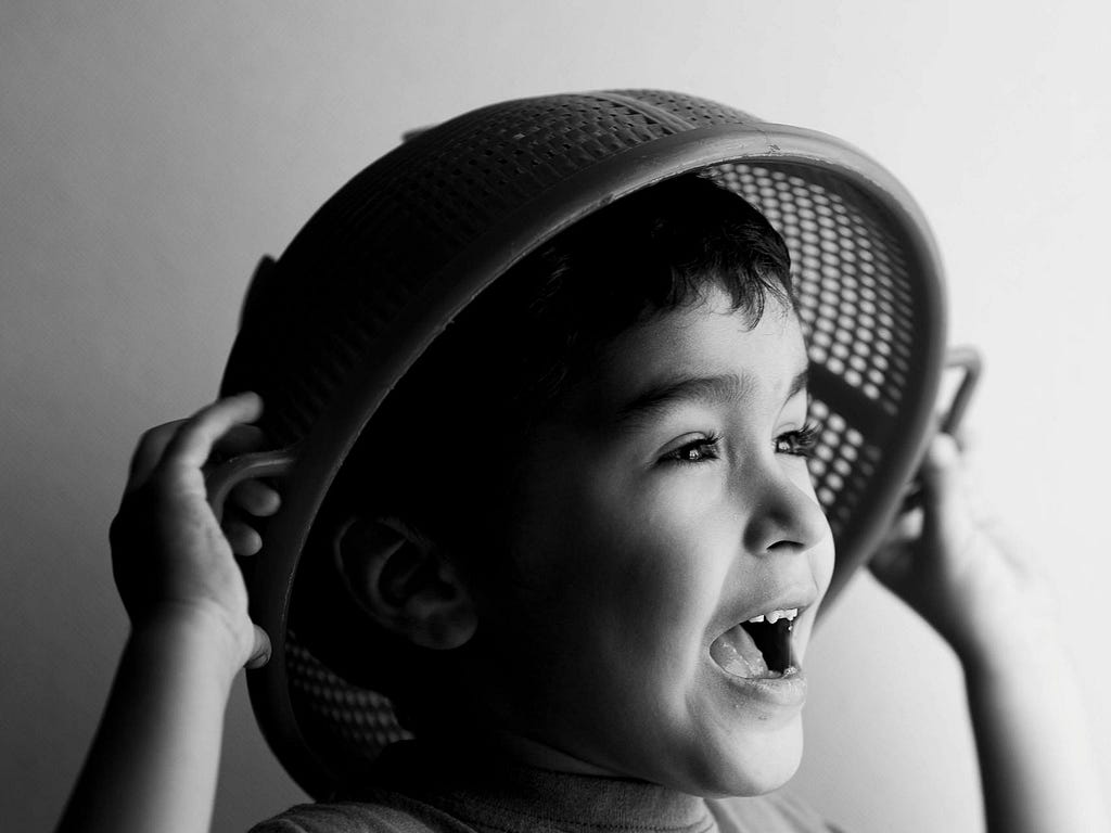 A greyscale photo of a boy holding a strainer over his head.