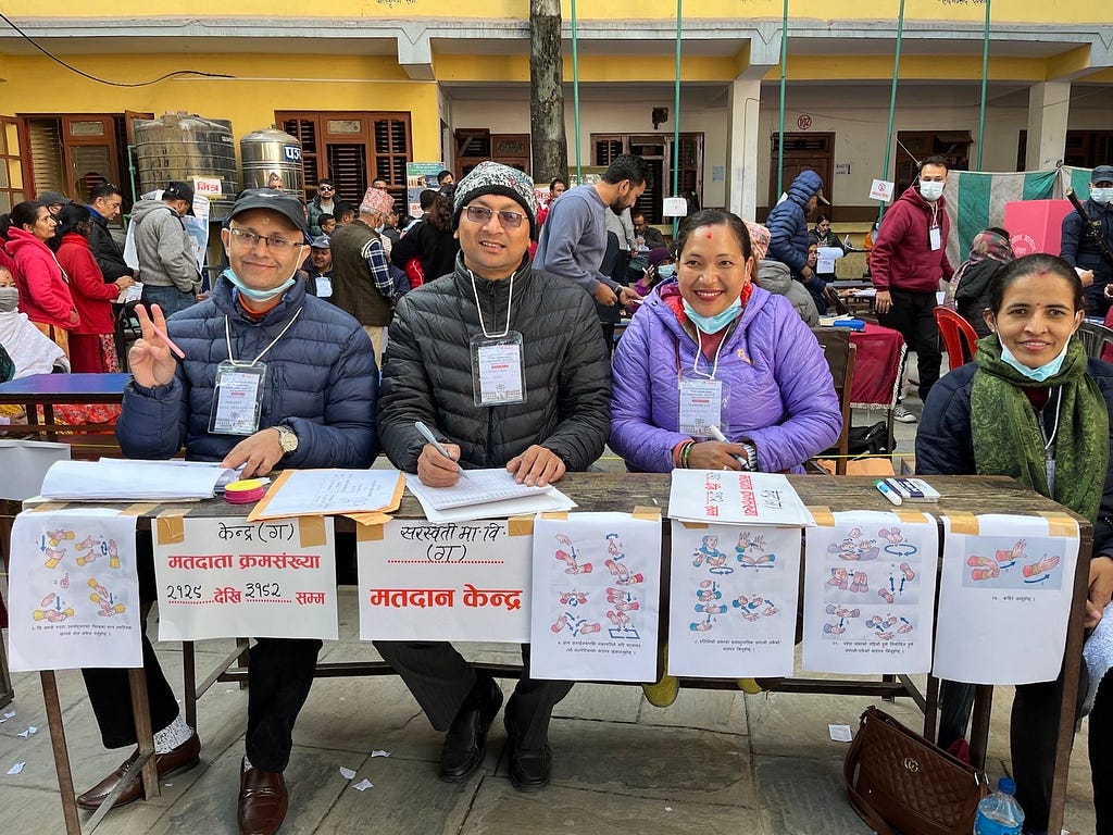 Four smiling people sit behind a polling station information table that includes several signs taped on the front that provide election instructions in the Nepali language.