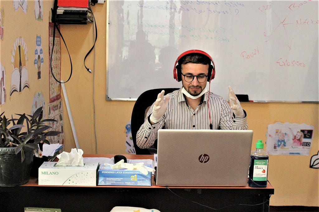 Man wearing a mask and gloves sits in a classroom in front of a laptop.