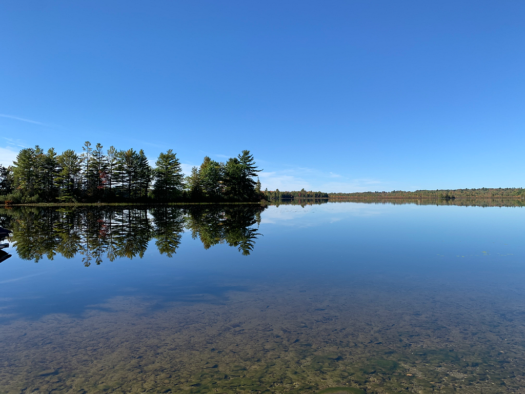 A picture of a calm and clear lake in Maine with the reflection of trees in the water.