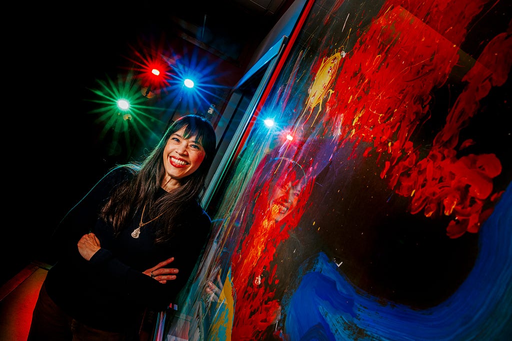 Karen Wills smiles for a photo in front of a painting in the UPC office with blue, green and red lights shining behind her