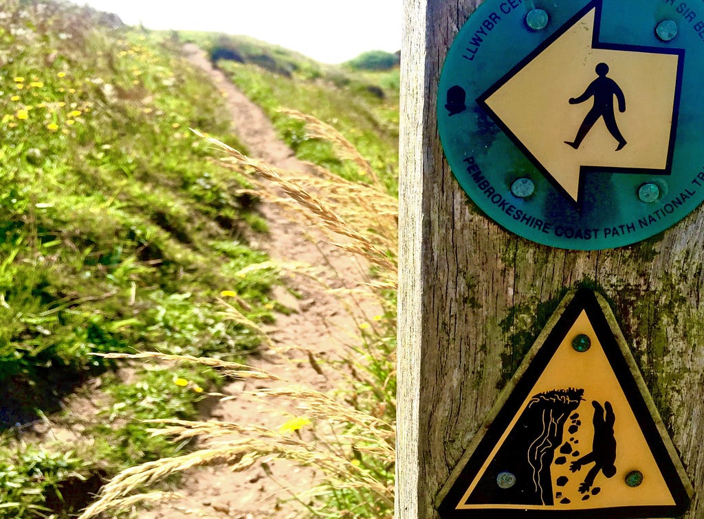 An uphill view of a winding path along a cliff edge. There are two signs in the foreground that give warning to walkers to keep to the path to avoid a dangerous fall.