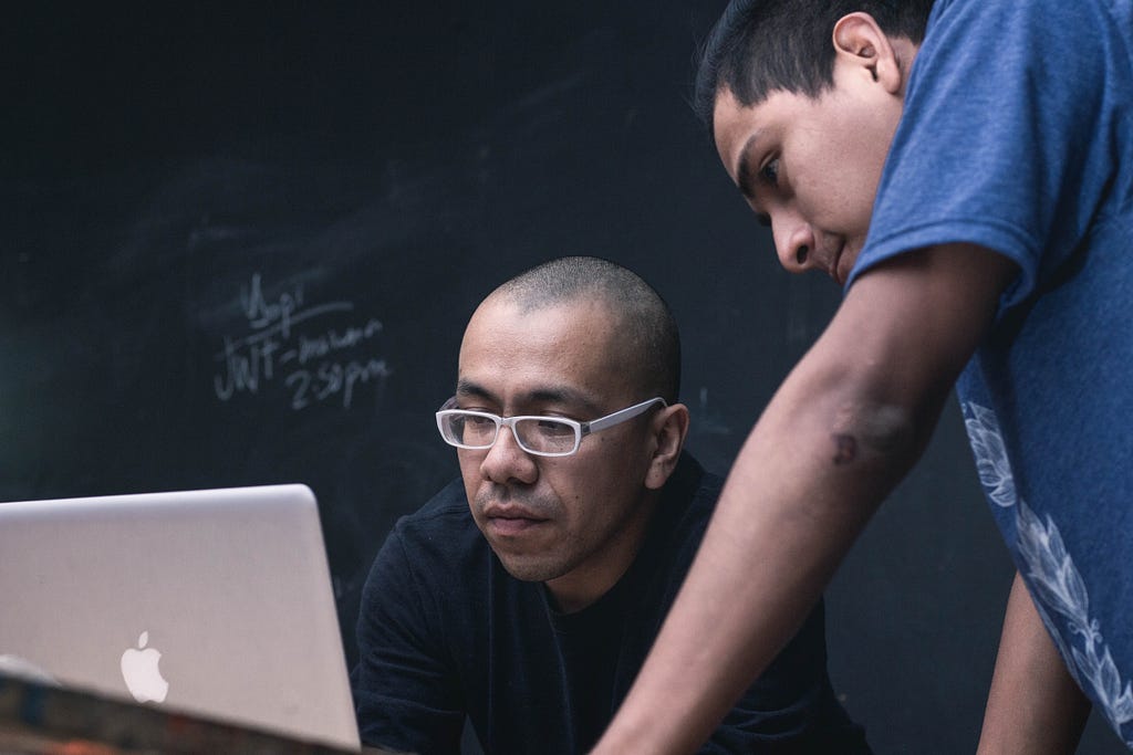 Man sitting looking at mac laptop with another man looking over his shoulder.