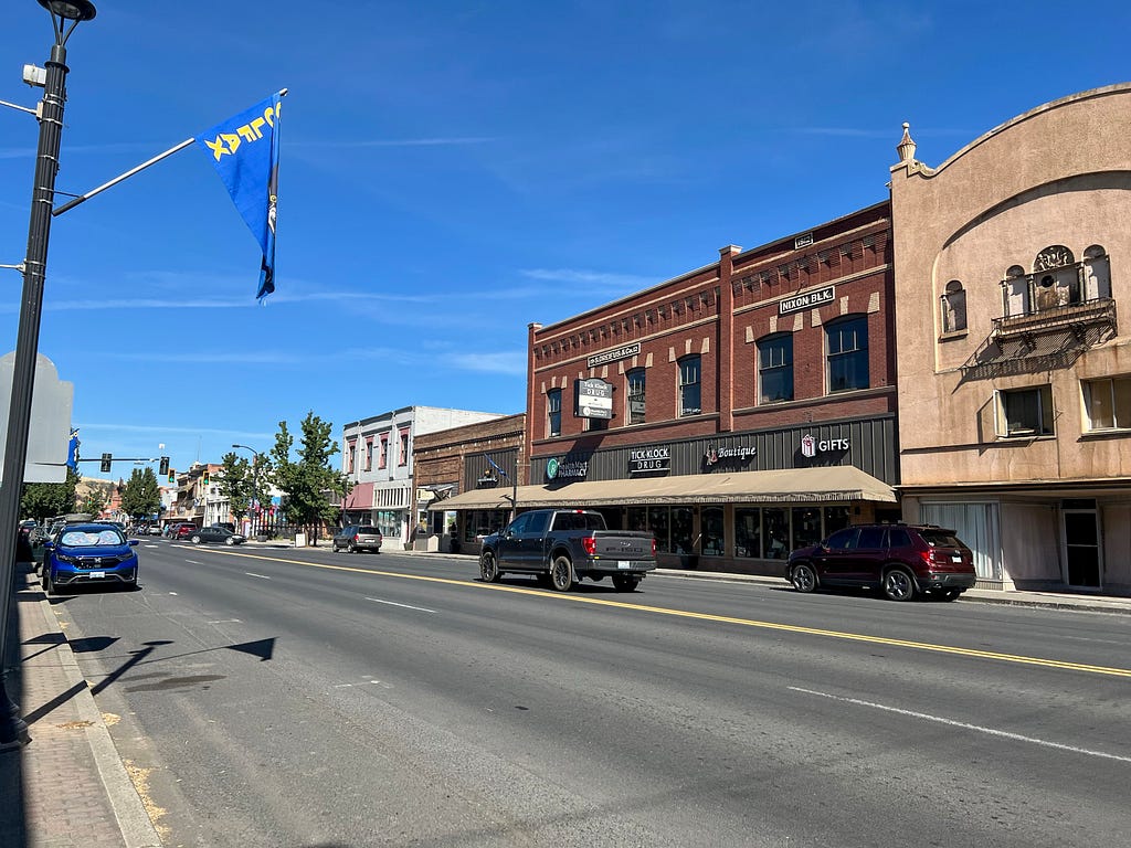 A row of historic buildings along a four lane street. A blue flag with yellow text reading “Colfax” frames the top against a blue, nearly cloudless sky. Cars are parked in front of buildings and one truck is visible driving further down the street.