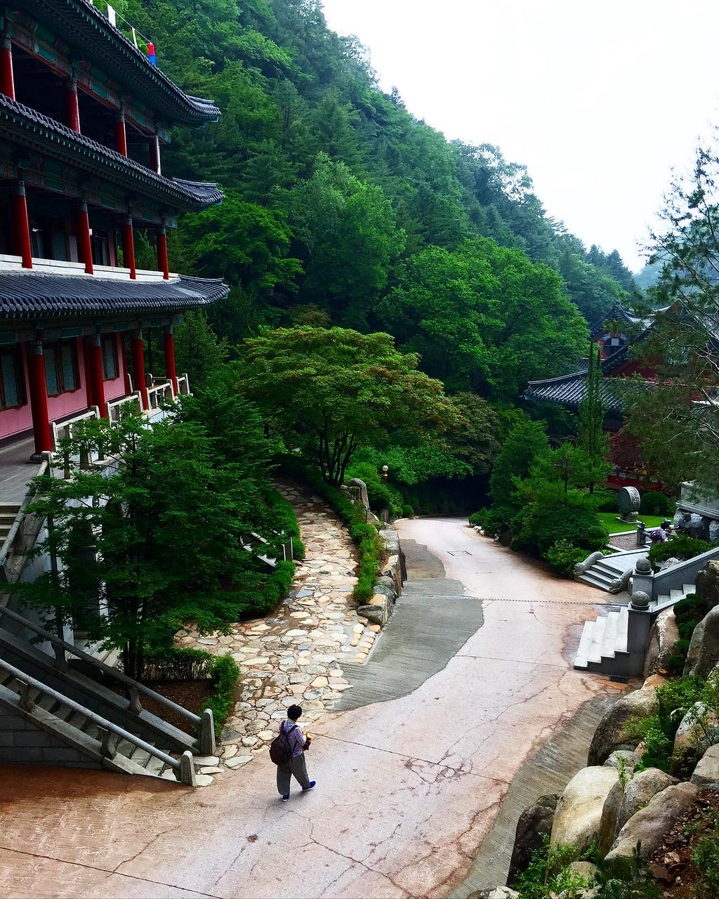 The bottom of the temple. A pathway towards the exit of the temple surrounded my luscious green trees and a monk walking in the path.