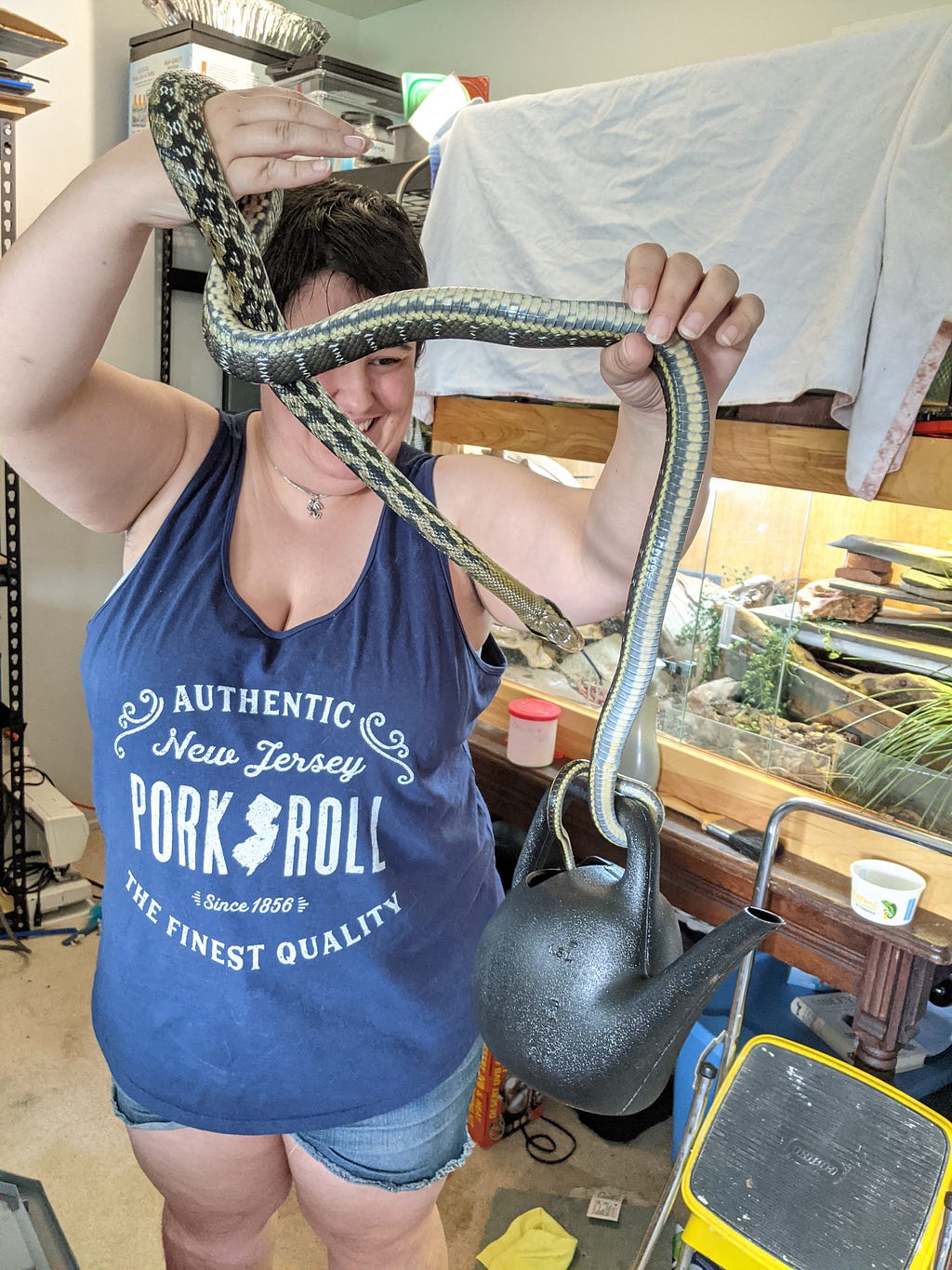 A short, chubby woman wearing an oversized blue tank top and denim shorts holds a long, slender snake up in front of her. The snake has a busy pattern comprised of diamonds fading to stripes in contrasting shades of dark and light green. The snake’s tail is wrapped around the handle of a small black plastic watering can, which it is holding well off the ground.