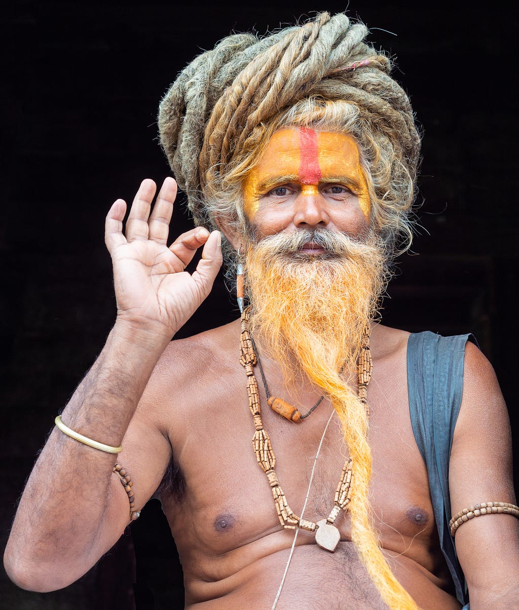 Image of a holy man with a long, twisted beard and intricate hairstyle, wearing traditional beads and paint on his face. He is making a hand gesture, creating a serene and spiritual atmosphere.