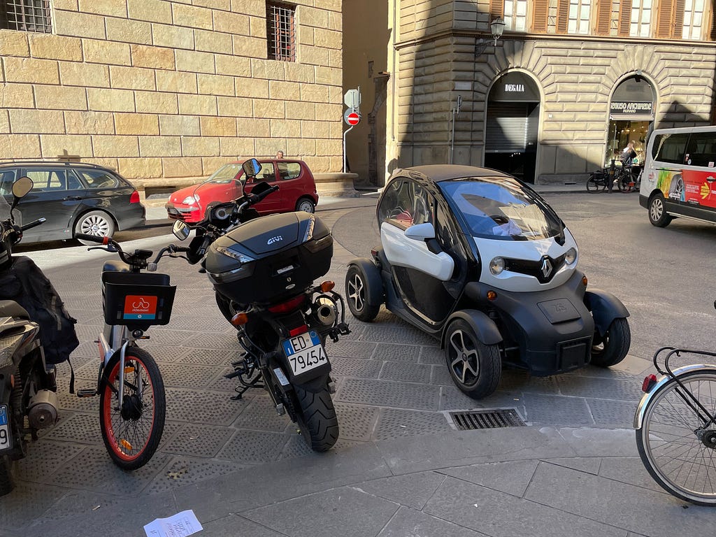 A bike hire, a motorcycle, and smart car parked next to each other.