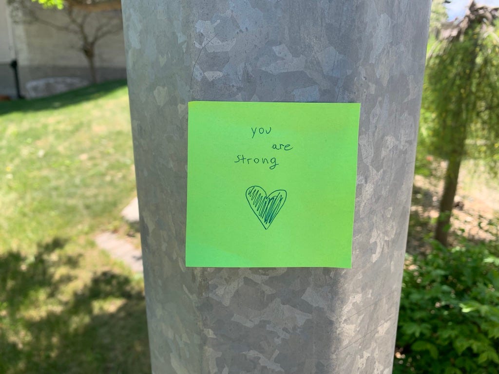 Lime green sticky note on a light post saying, “You are strong.”