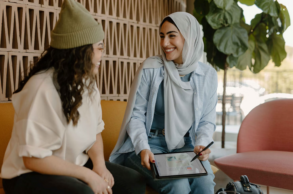 Two people sitting on a yellow couch, smiling at each other. One is dressed in a green beanie, white blouse, and black pants. The other is dressed in a grey head scarf, blue blazer, blue blouse, and blue jeans. They are holding an electronic tablet with a stylus pen.