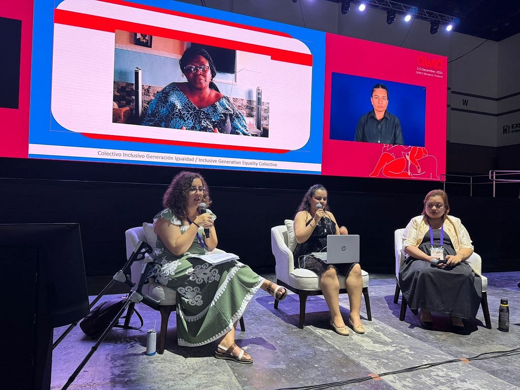 La autora comparte el escenario dentro del Forum AWID con otras dos mujeres, todas están sentadas y compartiendo con una audiencia que no sale en la imgen.