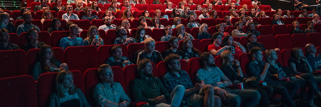 A crowd sitting in a movie theatre with red fabric seats.