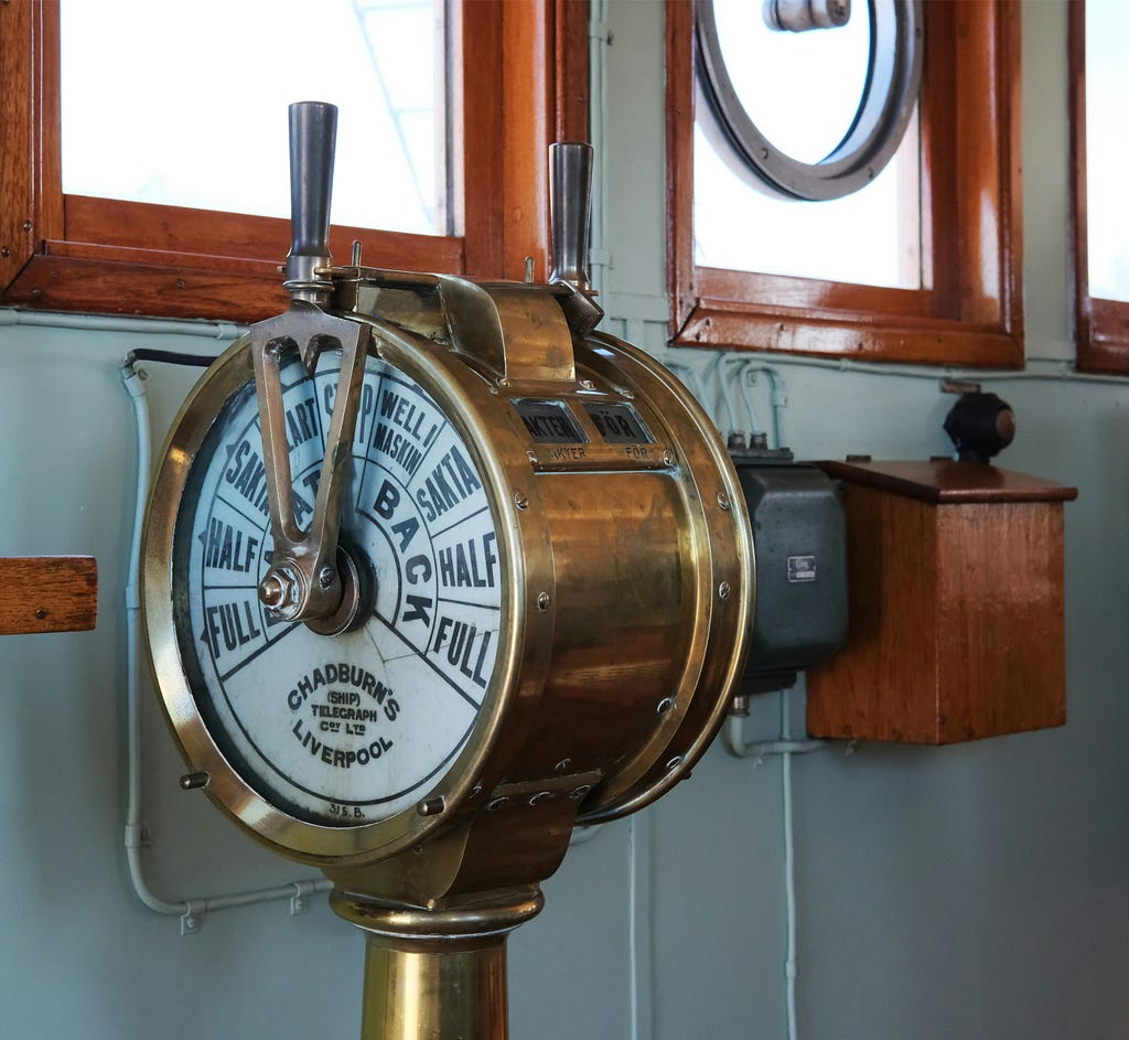 A brass ship’s engine order telegraph (ship’s telegraph) mounted on a pedestal in what appears to be a ship’s bridge or wheelhouse. The circular brass instrument has a white face with black markings showing different speed commands including “FULL,” “HALF,” “SLOW,” and “BACK.
