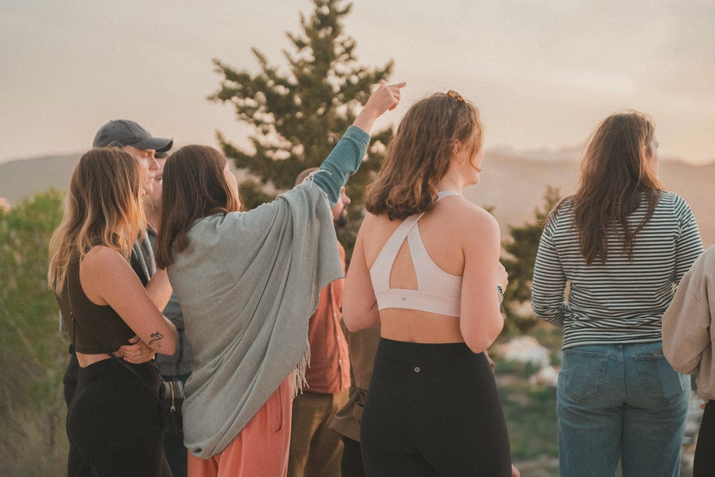 group of people standing togehter in nature with thier backs to the camera and one woman pointing at something in the distance