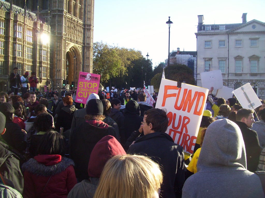 Photograph of students protesting in Westminster, 10 November 2010. Crowd with placards, seen from the back. In the distance the Houses of Parliament, sun reflecting in a window.