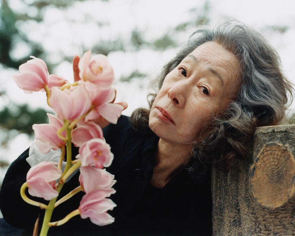 Na foto, vemos a atriz Yuh-Jung Youn com a cabeça apoiada em um tronco de árvore cortado. Ela tem uma expressão serena e segura flores cor-de-rosa.
