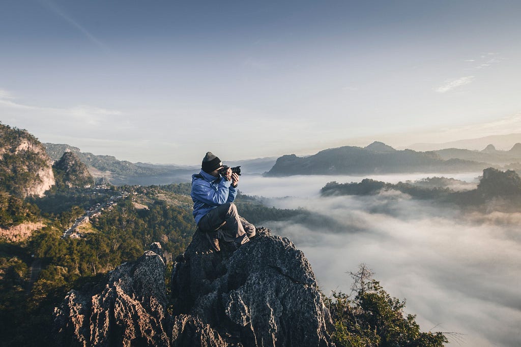 A photographer wearing a hat and blue waterproof looking through their camera while sitting on top of a mountain surrounded by clouds