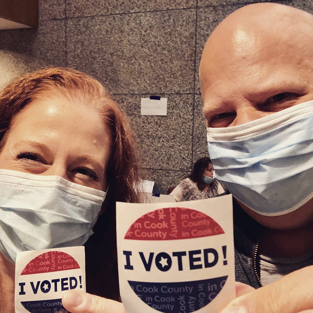 Photo of the author and her husband, wearing masks, and holding up “I Voted” stickers.