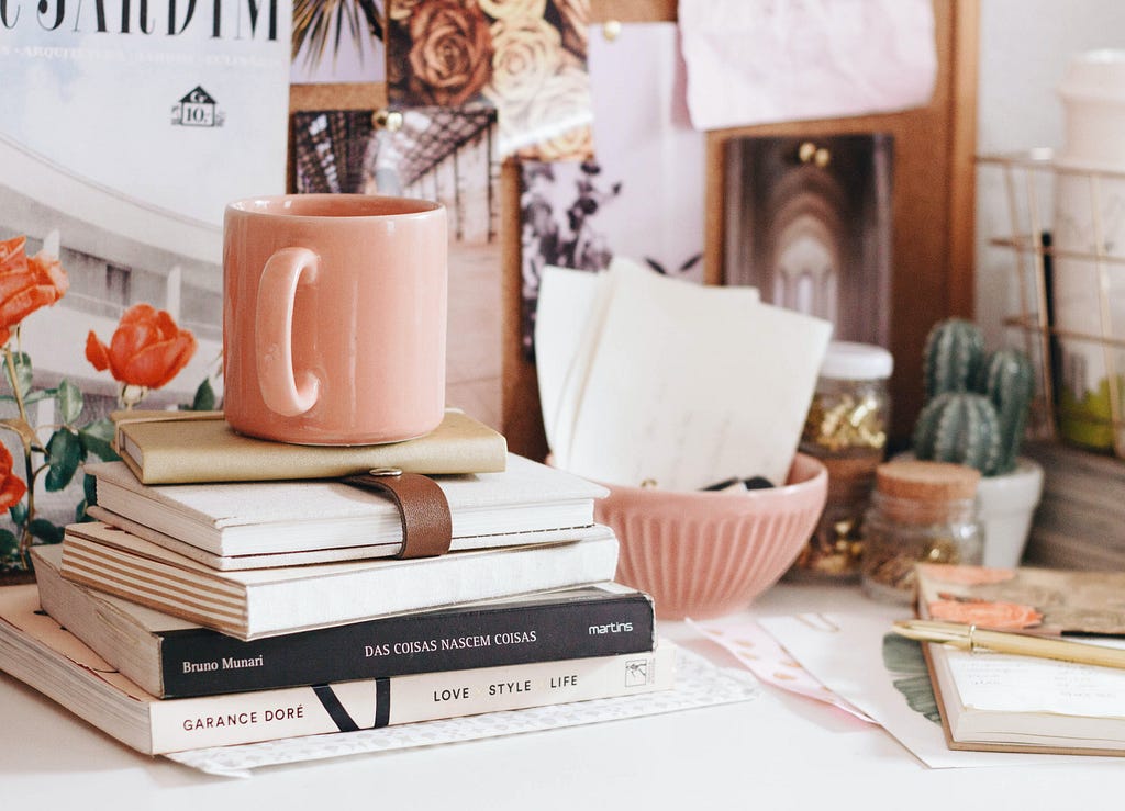 a cluttered desk, with a pastel pink mug resting on top of a pils of books. theres a potted cactus in the background and a few print outs pinned in the back splash.