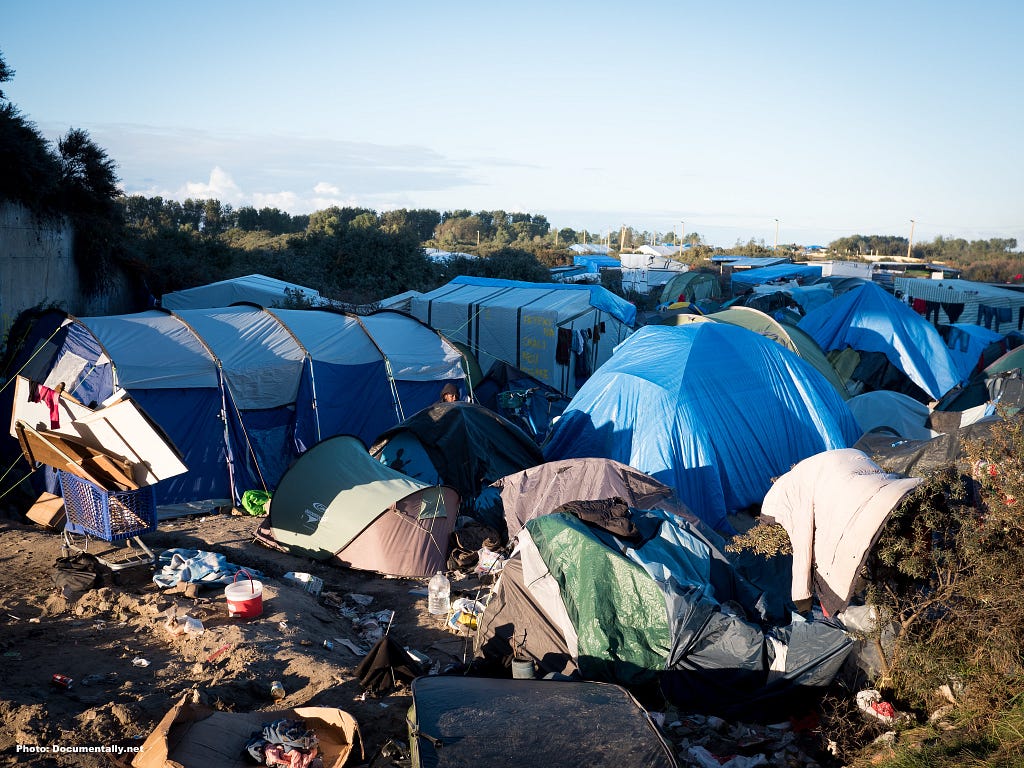 The scene is of an untidy collection of blue tents. There are some makeshift shelters in the foreground and some rubbish on the ground. The sky is blue and, in the chaos, it looks peaceful. In the distance, there are some trees. An image  taken by Christian Payne AKA Documentally. We have the Calais migrant camp in France.