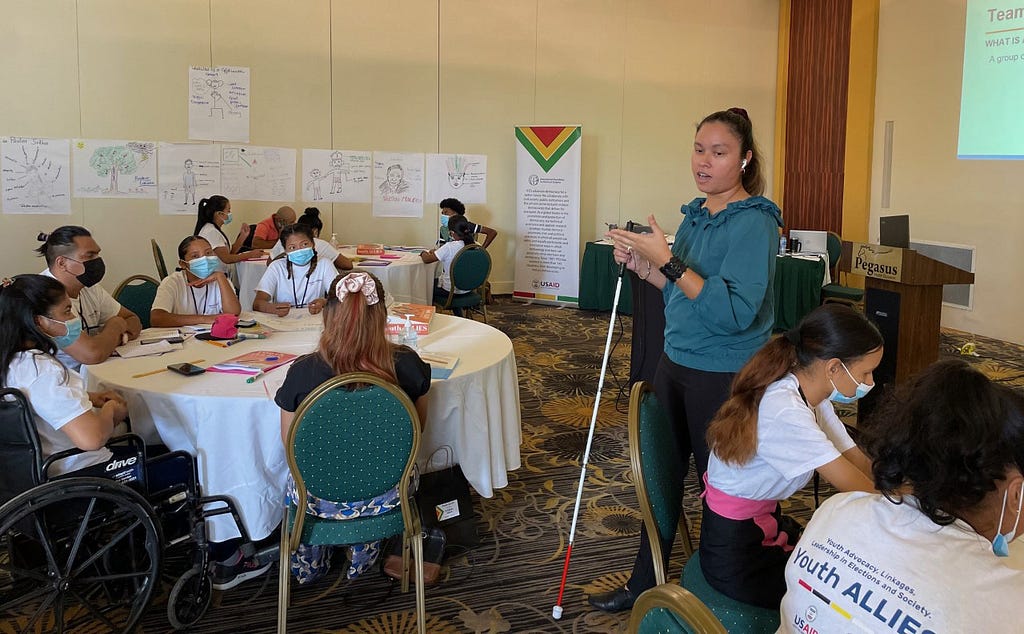 A young woman holding a white cane speaks to groups of people sitting at circular conference tables. The walls of the conference room are covered in notes taken on large sheets of white paper.