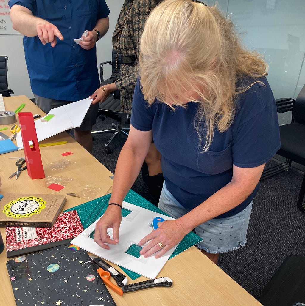 Photo of a teacher who is leaning over a table to work on  a craft project on cutting mat. She has 3 notebooks surrounding her. 2 teachers are in the background, pointing at her project.