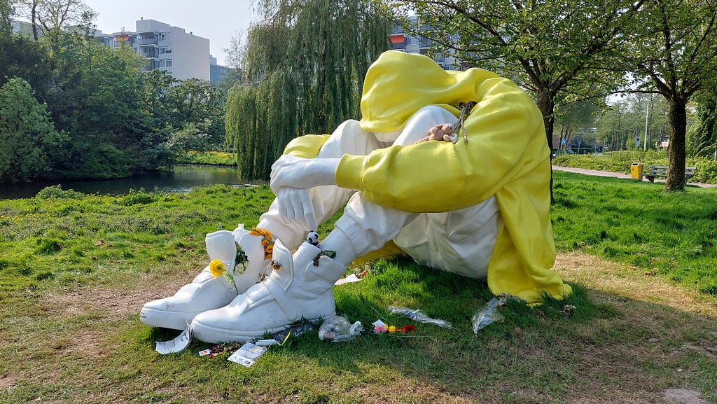 Image shows a huge statue in a park of a young person sitting with their legs pulled up, and their arms around their knees. The person wears a yellow jacket, white pants and white sneakers.