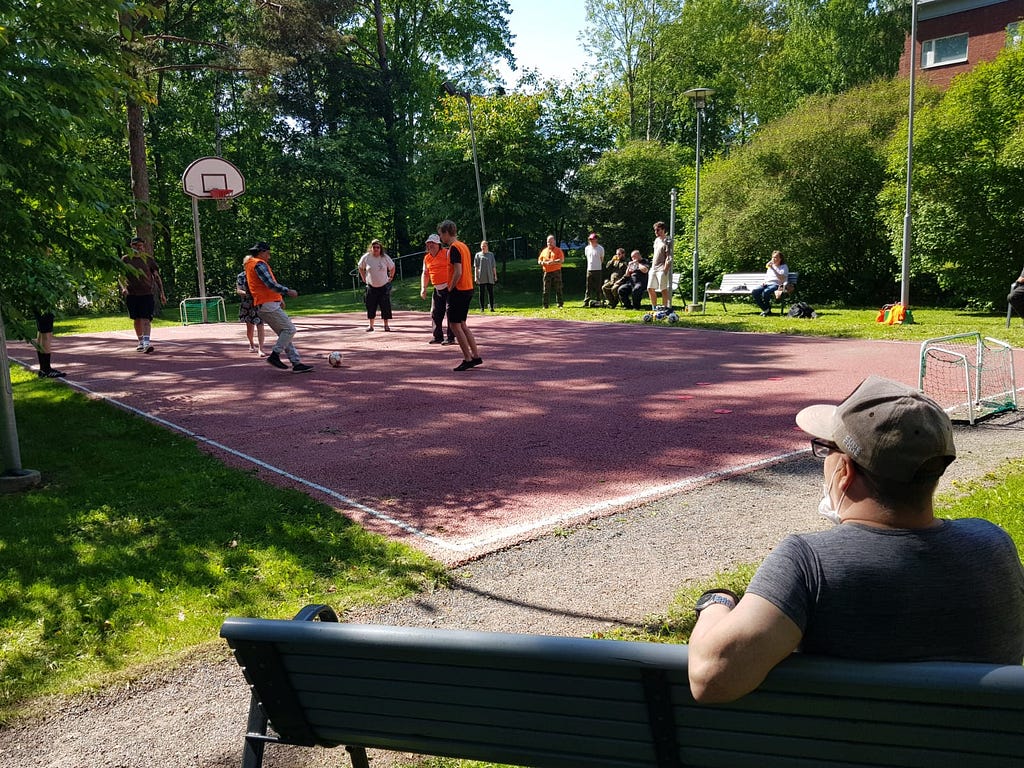 A group of people playing “walking football” on a red sports court outdoors while a spectator looks on