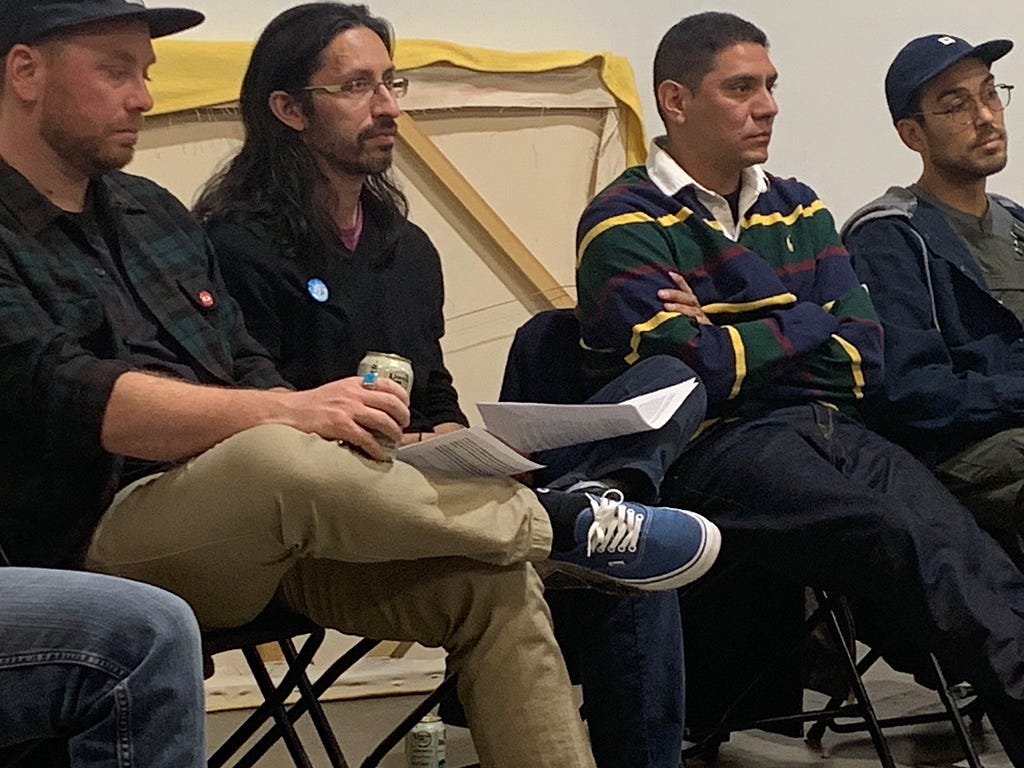 Four men sit listening. On the far left is a white-appearing man wearing khaki pants and a black shirt and blue sneakers. Next to him are three Latinx-appearing men: one with long hair and glasses, one with short hair wearing a rugby shirt, and one with a baseball cap and glasses.