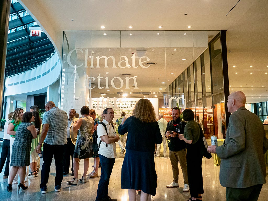 A group of people are at a party hosted by the Climate Action Museum, with the large letters of the museum’s name on a glass wall behind the group.