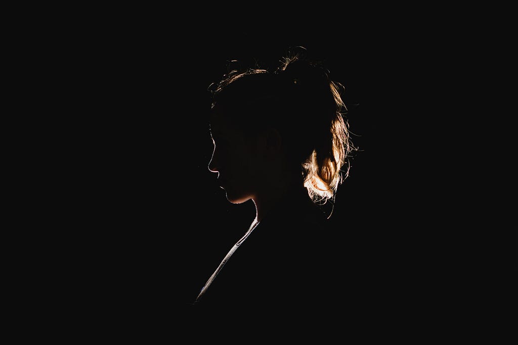 A female figure, from the shoulders up faces to the left in profile. A black background and backlit, so only small details and hair can be distinguished.