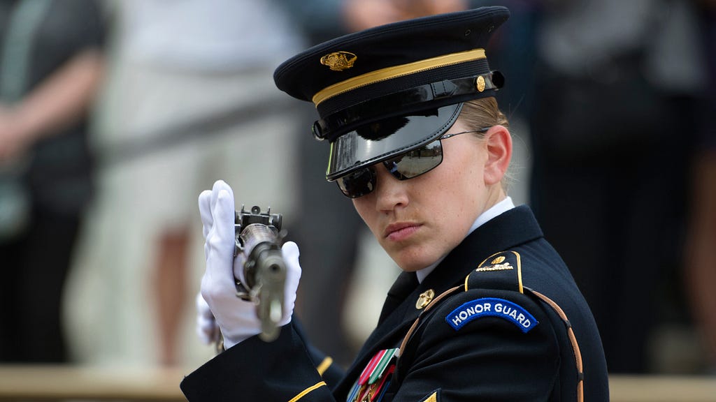 A woman in sunglasses, gloves, and an honor guard uniform looks down at a rifle. The barrel of the gun faces the camera.