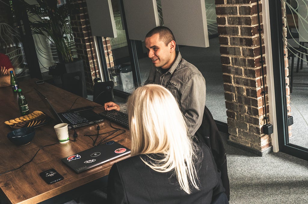 Two people sitting by the desk. The man — Przemek, is in front of the computer testing software. The woman — Ania,  is sitting and listening to him speaking.