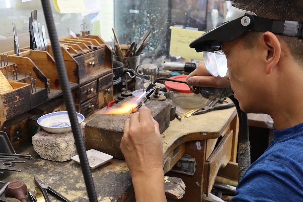 Phillip welds a ring with a blow torch on top of a rectangular slab that sits on his work bench. His head and shoulders takes up the right quarter of the frame. He wears safety goggles and a blue t-shirt. Behind the slab on the desk are small wooden drawers and containers for intricate jewelry making tools.