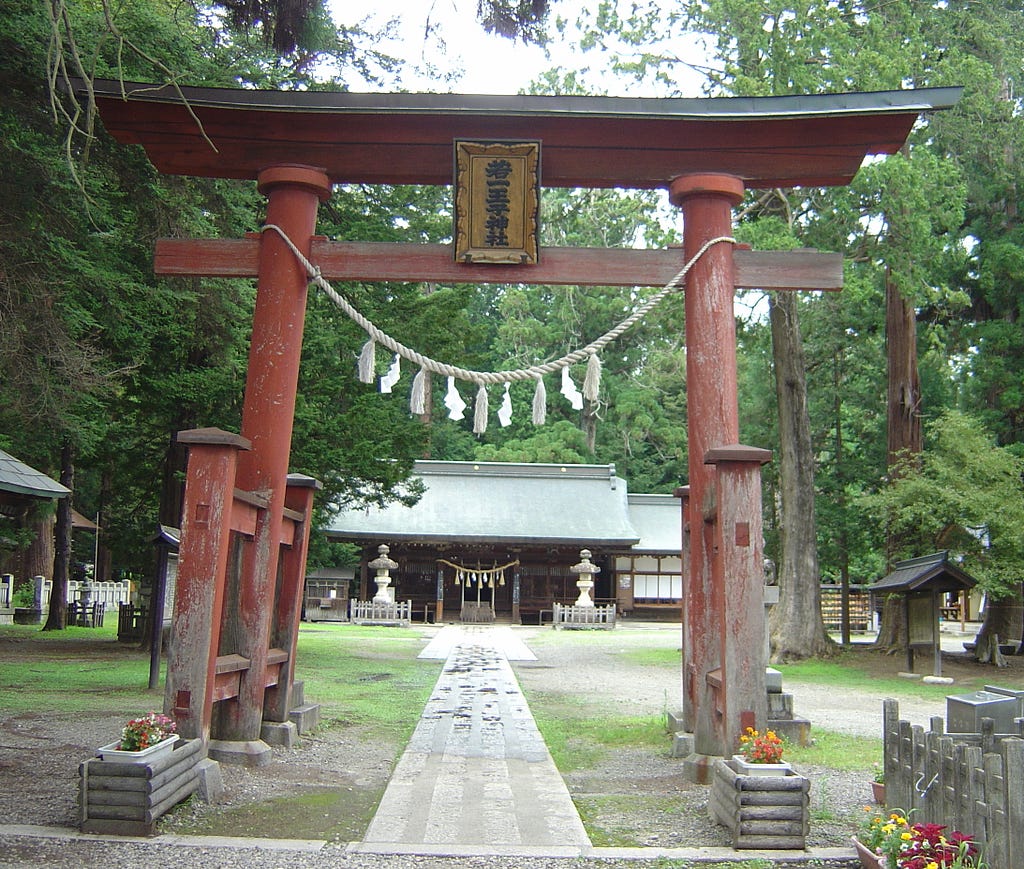 A weathered tori gate stands in a clearing surrounded by trees. A rope with tassels and folded paper gohei hangs from it. Beyond it, at the end of a stone path, is the shrine building. The offering box and entrance in the previous photo are just visible. Large stone lanterns stand on either side of the entrance.
