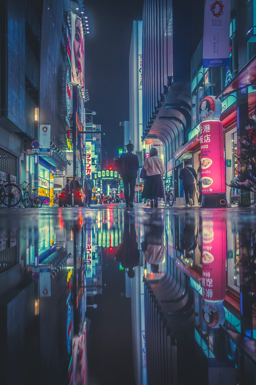 Two people walk down a colorfully lit city street. It appears as though it has just rained. The lights of the city reflect off of the glassy ground, giving the image a magical quality.